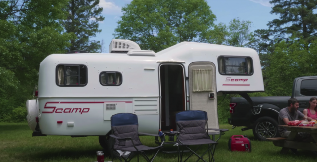 Photo of a Scamp trailer in a green outdoor setting with people and camping chairs